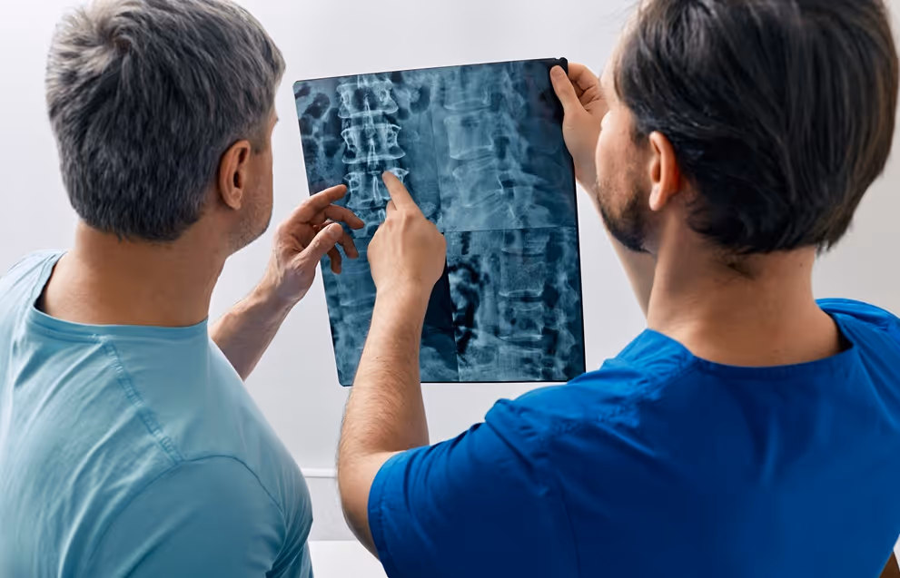 Two men examine a spinal X-ray, one pointing at the vertebrae while discussing the image.