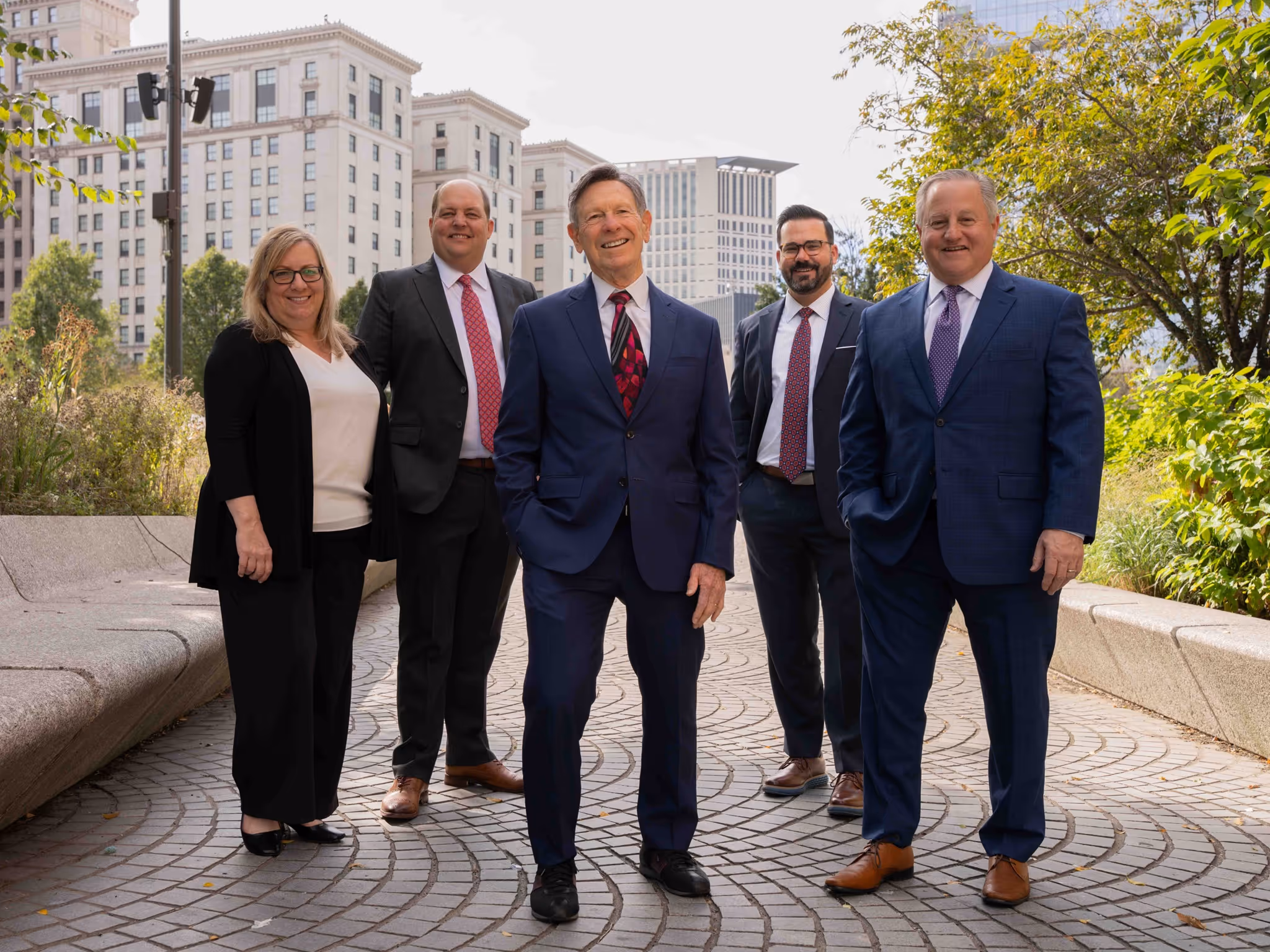 Group of five business professionals standing outdoors on a paved area with buildings and greenery behind them.