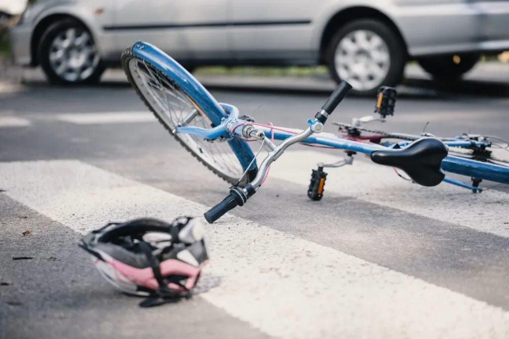 Blue bicycle lying on a crosswalk with a helmet on the ground and a car in the background.