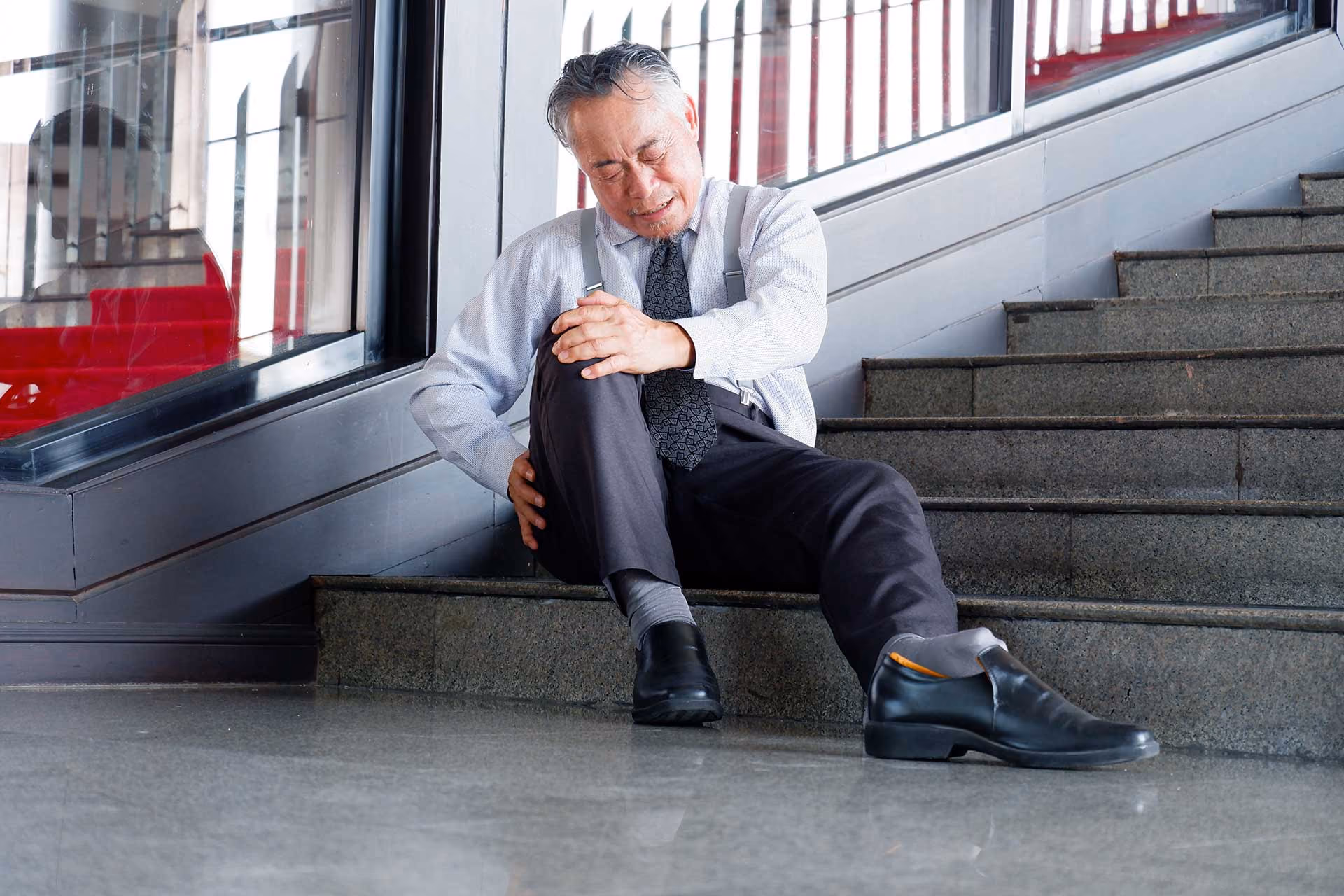 Man in business attire sitting on stairs holding his knee in pain with eyes closed.