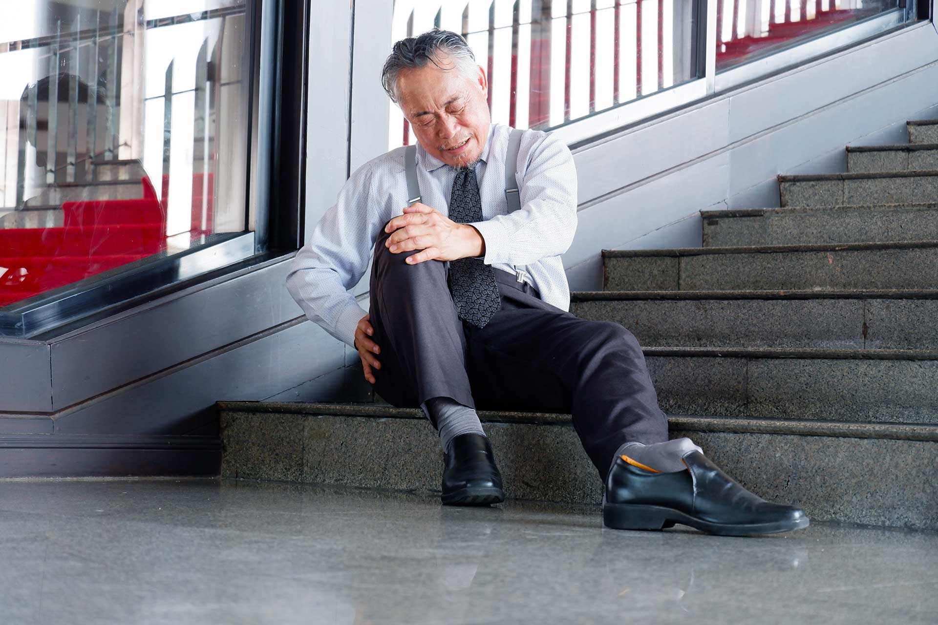 Man in business attire sitting on stairs holding his knee in pain with eyes closed.
