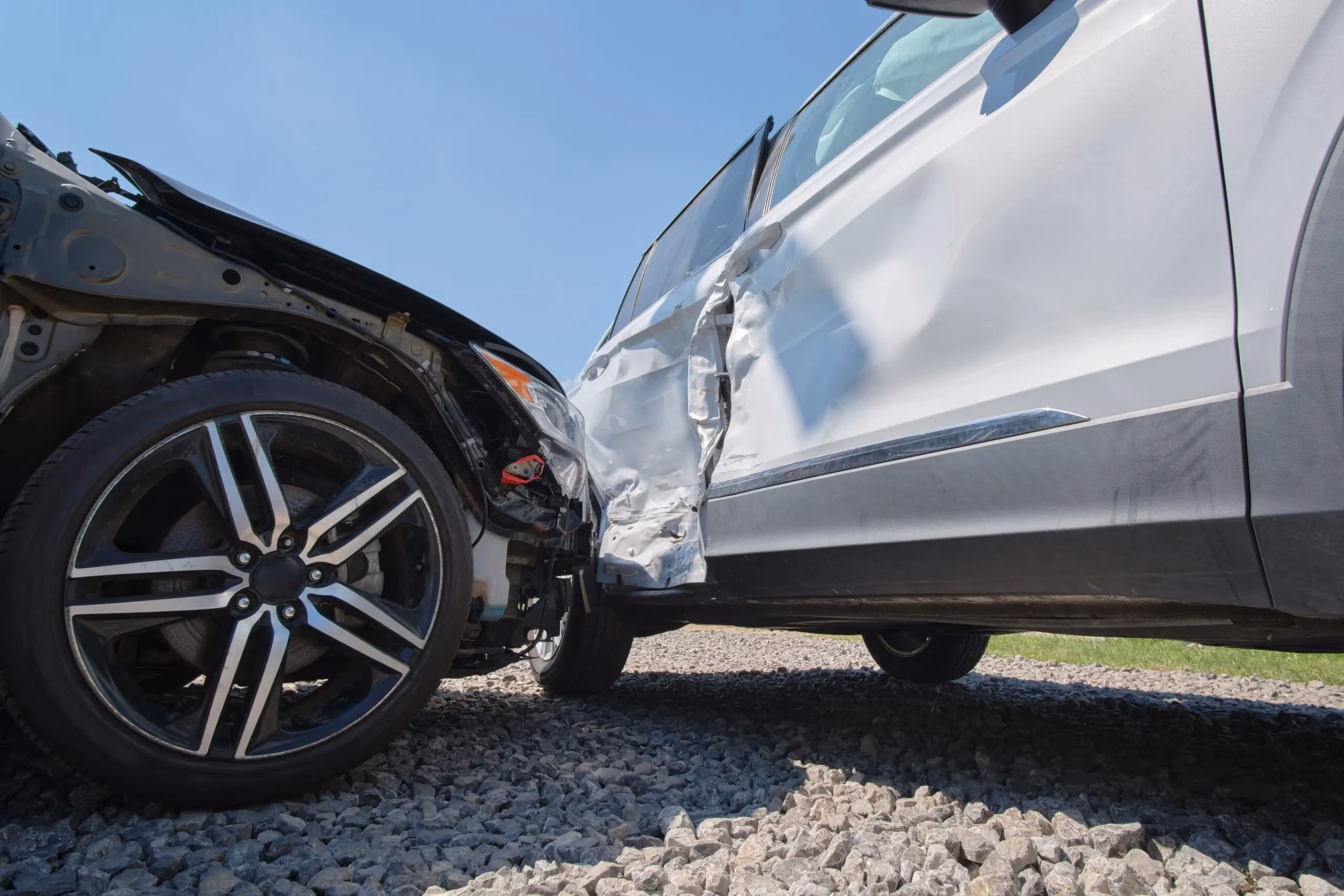 Two cars collide on gravel with significant damage to the black car's front and white car's side.