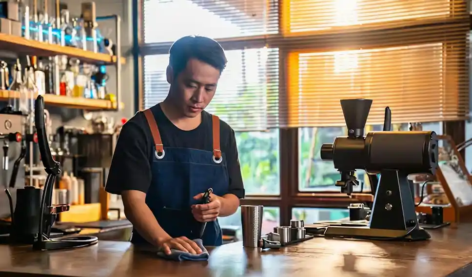 Barista in apron cleaning the counter in a sunlit coffee shop with espresso machine and tools nearby.