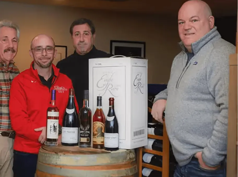 Four men standing behind barrels with several bottles of wine and a white wine box.