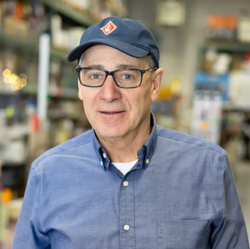 Older man wearing glasses and a blue cap and shirt, standing in a blurred store background.
