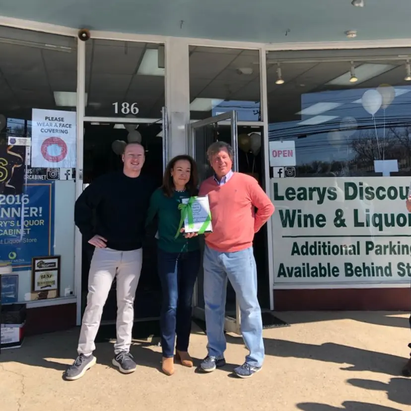 Three people standing outside Learys Discount Wine & Liquor store holding an award plaque with a green ribbon.