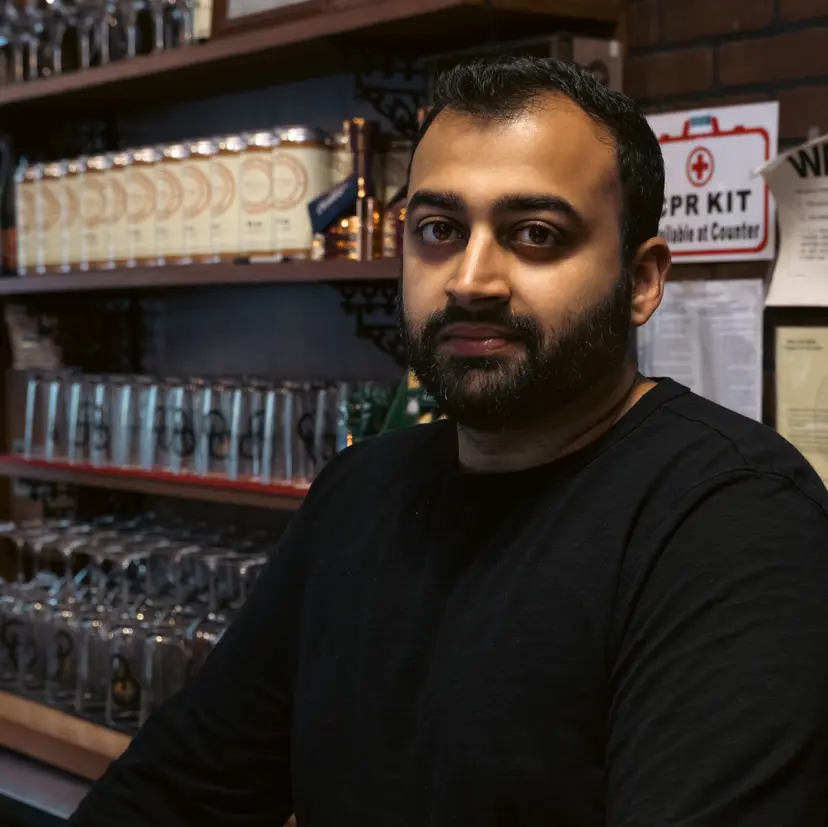 Bearded man in black shirt stands in front of shelves with bottles and glassware in dimly lit bar.
