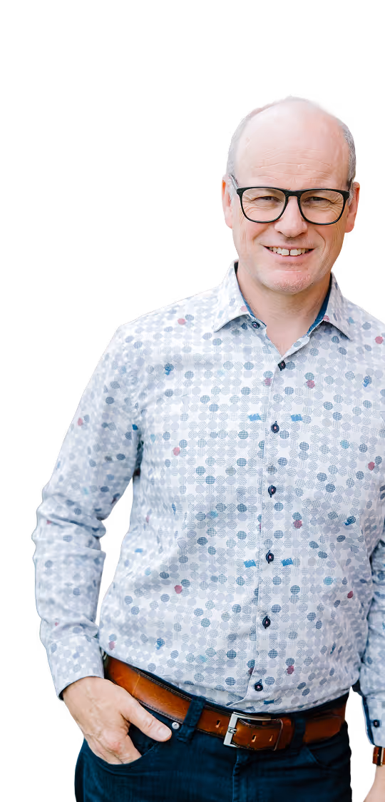Smiling middle-aged man wearing glasses, patterned shirt, brown belt, and dark pants, hand in pocket.