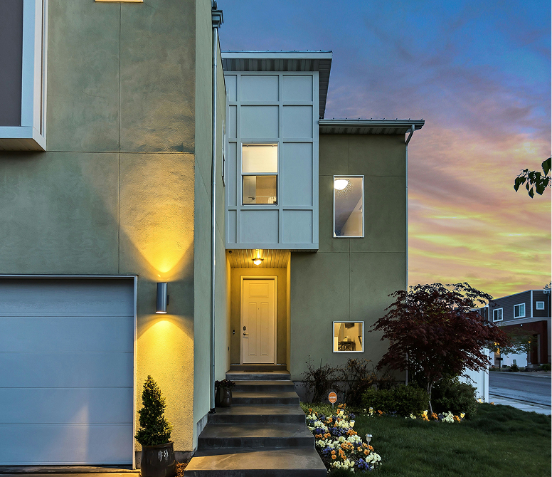 Front porch with lit wall sconces, blue door, wooden steps, and various potted plants at dusk.