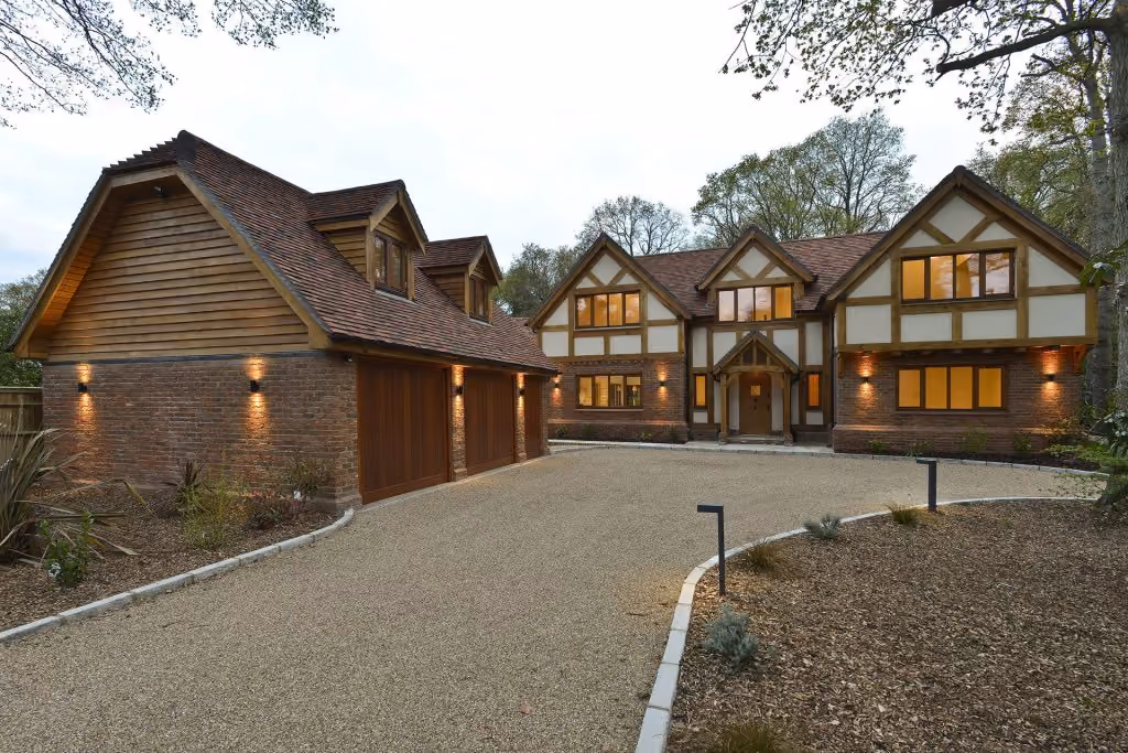 Large two-story house with brick and timber exterior, lit windows, and a three-car garage on a gravel driveway.
