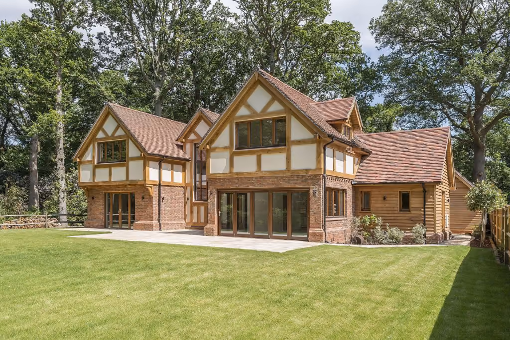 Large two-story Tudor-style house with brick, wood, and large windows surrounded by green lawn and trees.
