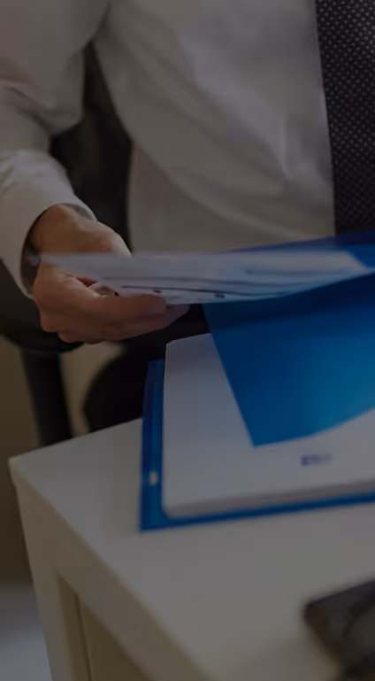 Person in white shirt and tie holding papers over a white desk with a blue folder on it.