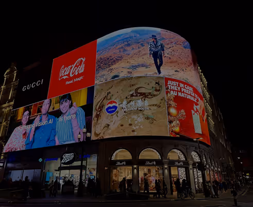 Night view of building with illuminated large digital billboards showing Coca-Cola, Gucci, Pepsi, and other ads.