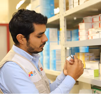Man in a vest examining a medication bottle in a pharmacy or medical supply storage room.