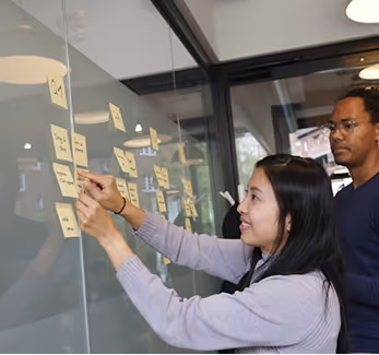 Two colleagues placing sticky notes on a glass wall during a collaborative work session.