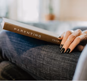 Person holding a book titled 'PARENTING' with black nail polish and rings on fingers.