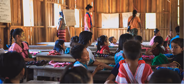 Children in colorful traditional clothing seated at wooden desks inside a wooden classroom with teachers.