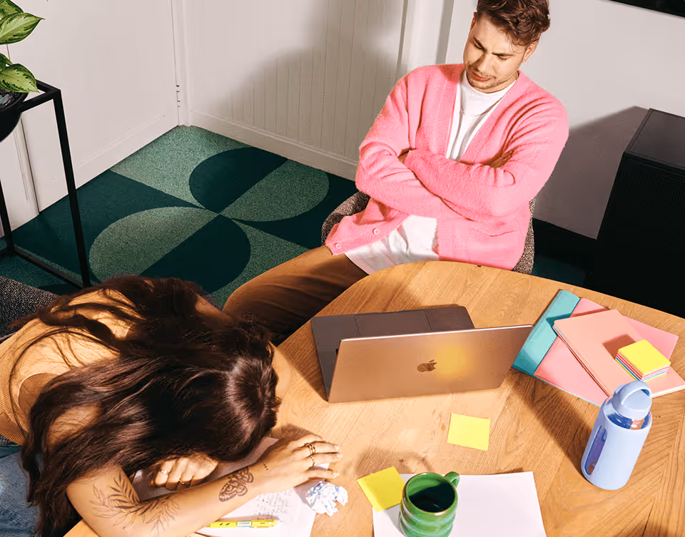 Woman resting her head on the table while a man in pink sweater watches, with laptop and notebooks on table.