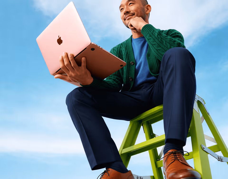 Man in green cardigan sitting on green ladder holding a rose gold Apple laptop against a blue sky.