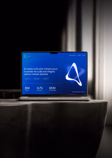 Open laptop on a table displaying a blue-themed webpage for AI-native verification infrastructure supporting carbon markets with financial metrics and a white triangular logo.
