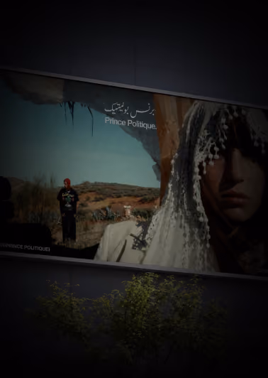 Outdoor billboard featuring a close-up of a person wearing a white lace veil with beaded details on the right side and another person standing in a desert landscape on the left side, with text reading 'Prince Politique' in English and Arabic.