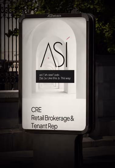Backlit advertisement sign on a street displaying the word 'ASÍ' with its pronunciation and meaning: 'así (ah-see) adv. [Sp.] a. Like this, b. This way'. Below, text reads: 'CRE Retail Brokerage & Tenant Rep', set against a background of white arches and a corridor.