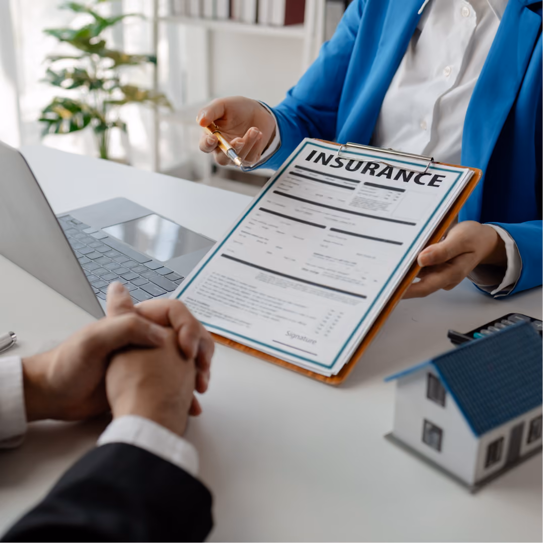 Insurance advisor explains policy details to a client across a desk with documents and laptop.