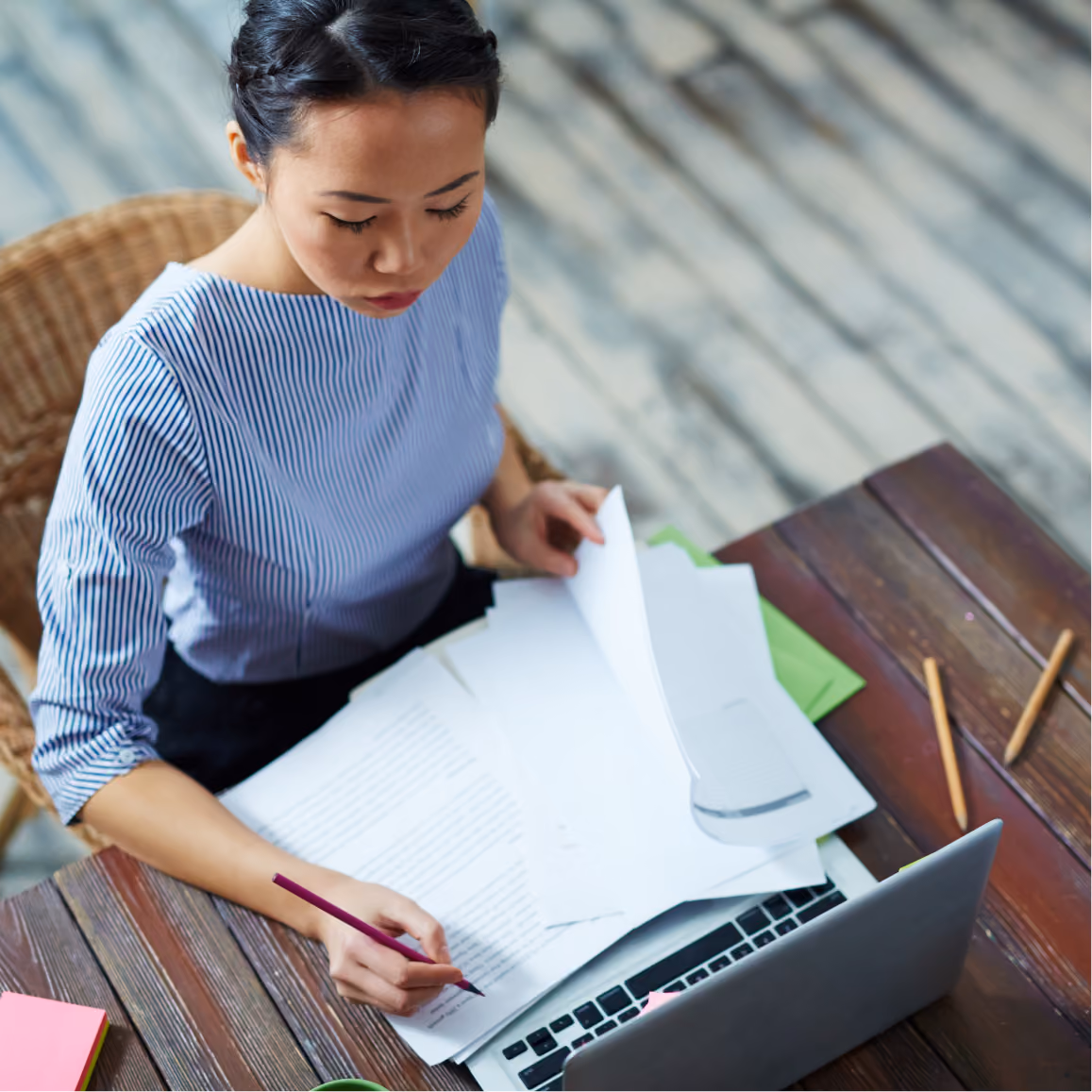 Professional reviews printed draft documents and makes notes beside a laptop at a wooden desk.