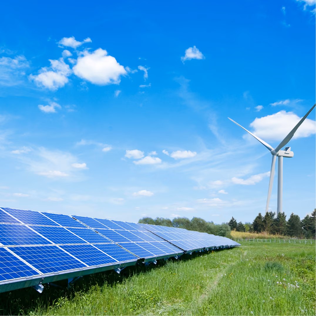 Rows of solar panels in a green field beside a tall wind turbine under a bright blue sky with scattered clouds.