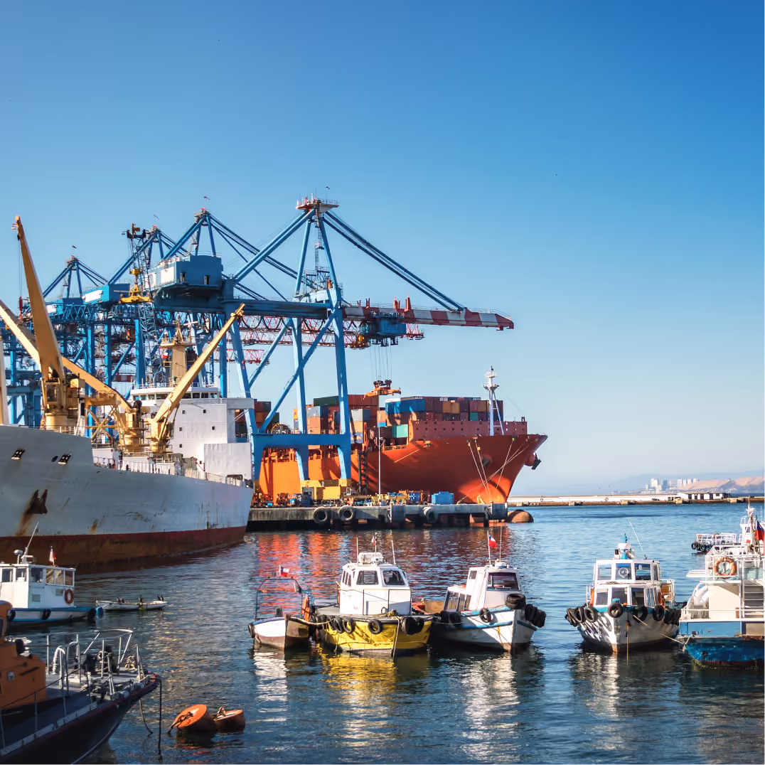 Cargo ship loaded with containers at a busy port, tall cranes above and small boats moored in calm blue water.