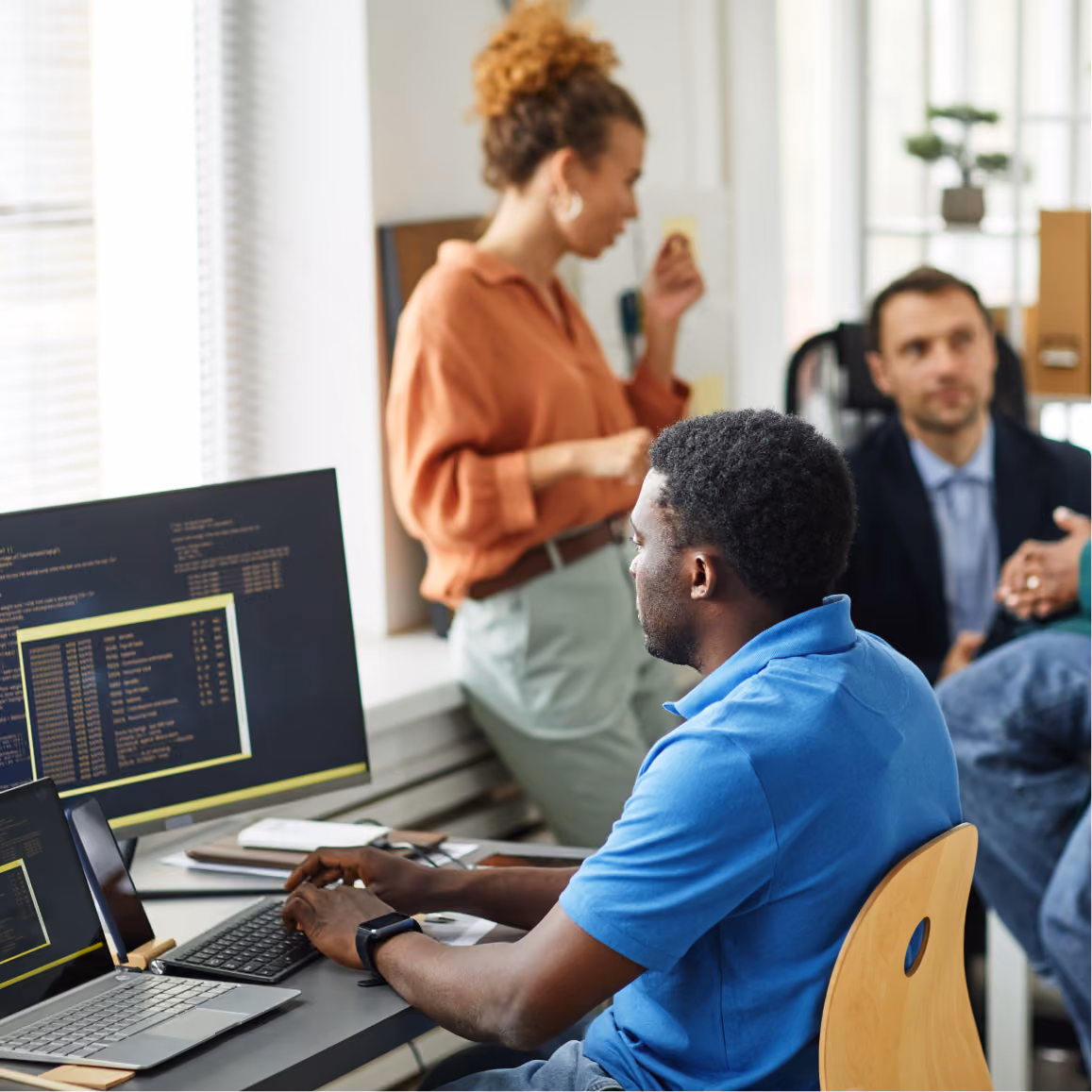 Software developer works at computer while teammates discuss project planning in collaborative office environm