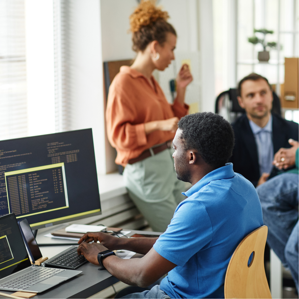 Software developer works at computer while teammates discuss project planning in collaborative office environm