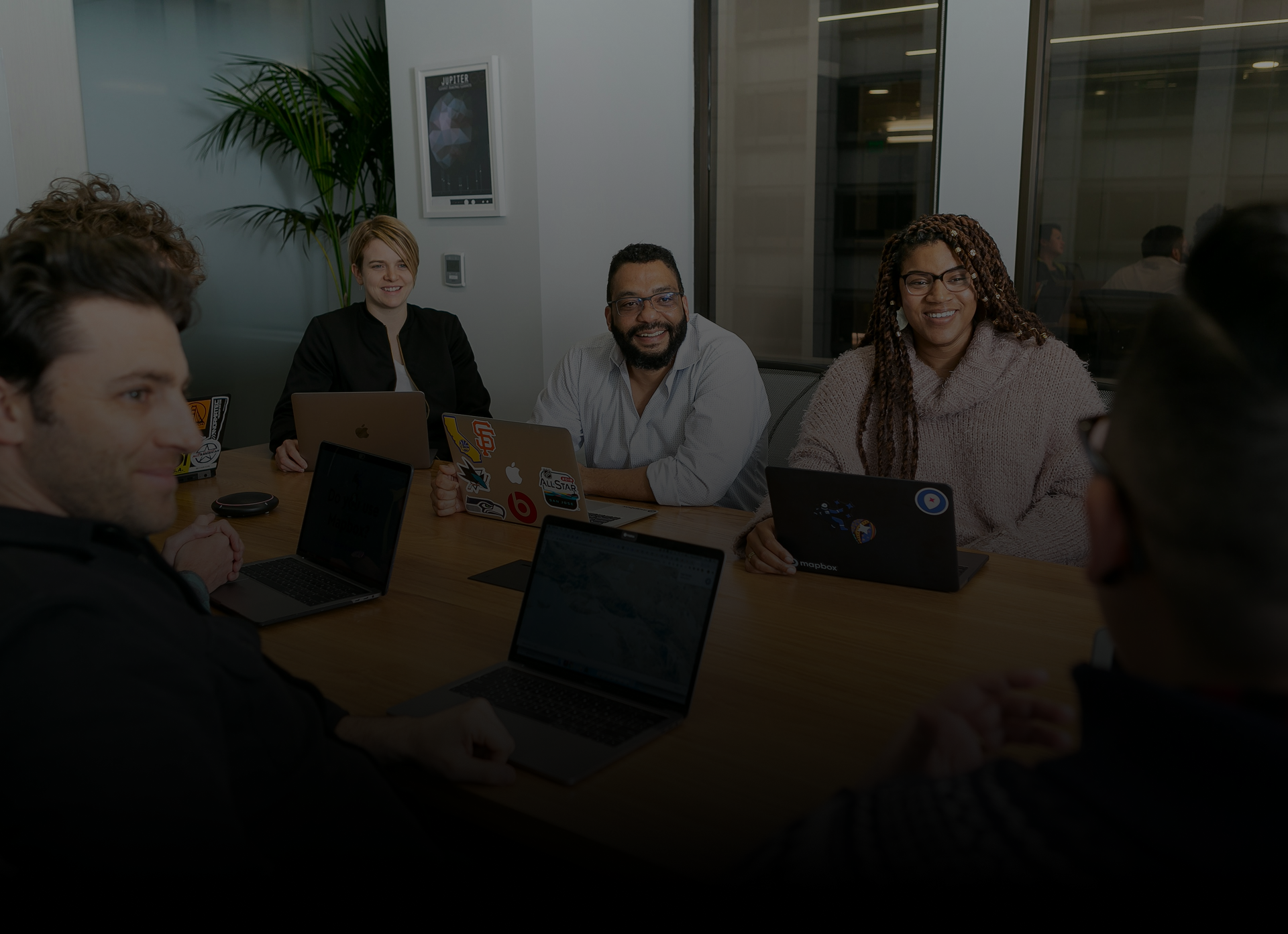 Five diverse colleagues seated around a conference table in an office, smiling and engaging in a meeting with open laptops in front of them.