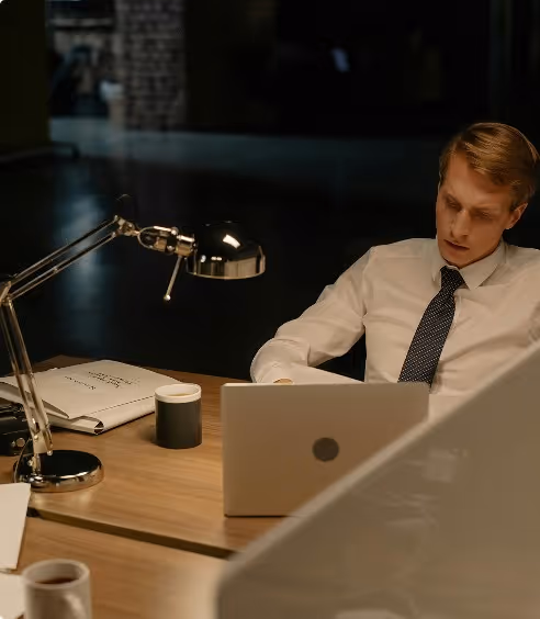Man in white shirt and tie working late at a wooden desk with a laptop and desk lamp.