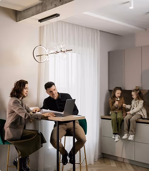 Two adults sitting at a table using a laptop while two children sit nearby on kitchen cabinets talking.