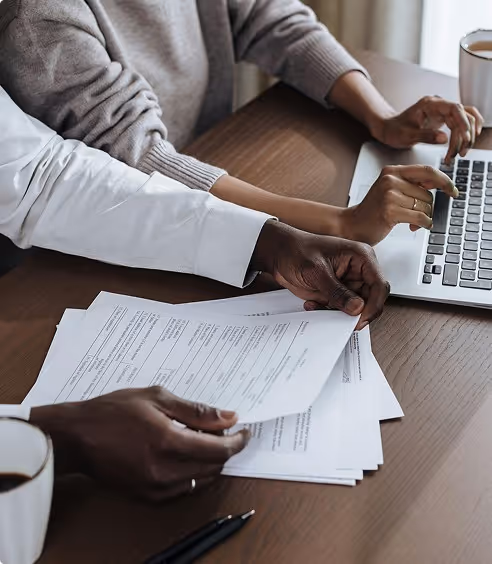 Two people reviewing papers and working on a laptop at a wooden table with coffee cups.