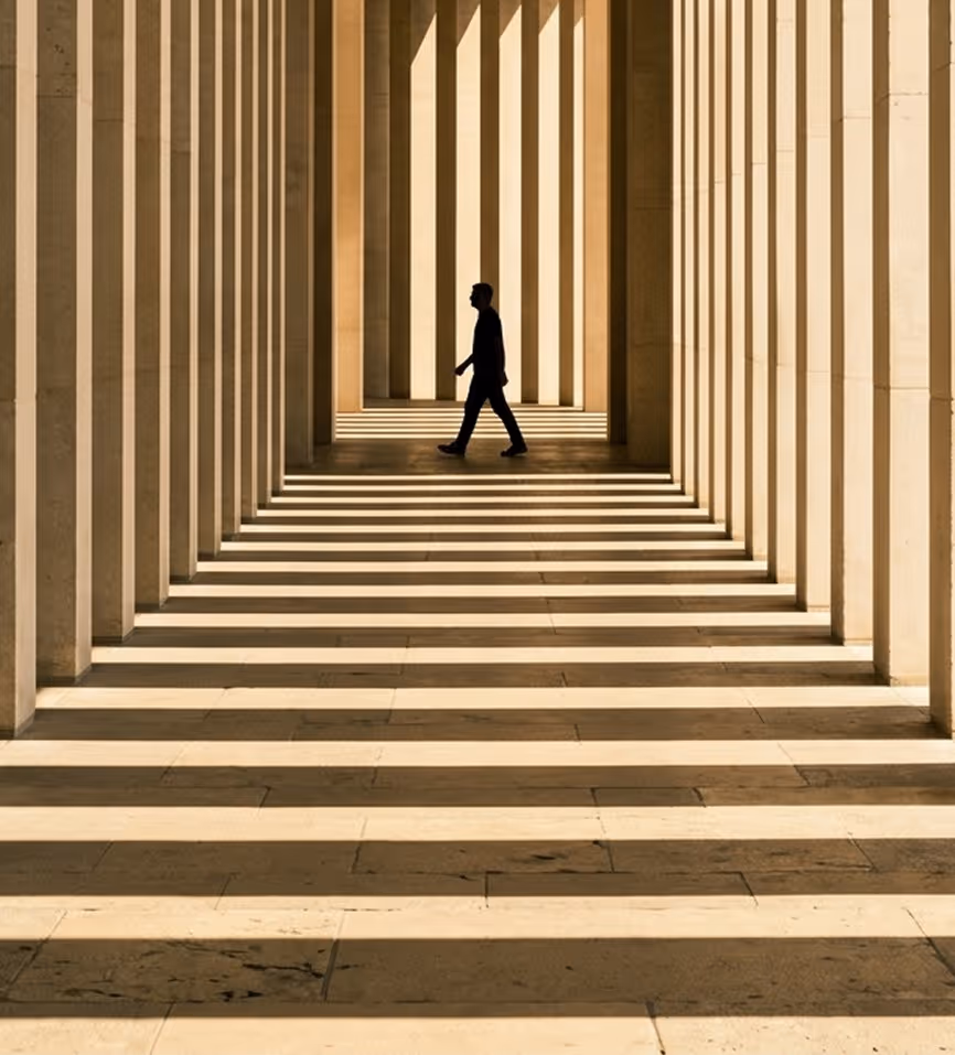 Silhouette of a person walking through a corridor with strong vertical and horizontal striped shadows.