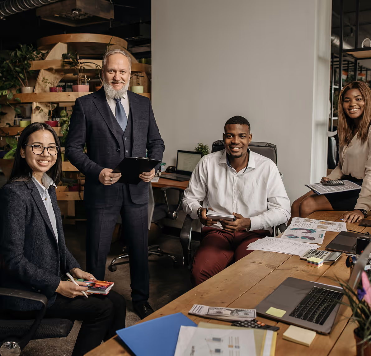 Four diverse business professionals smiling in an office around a table with documents and laptops.
