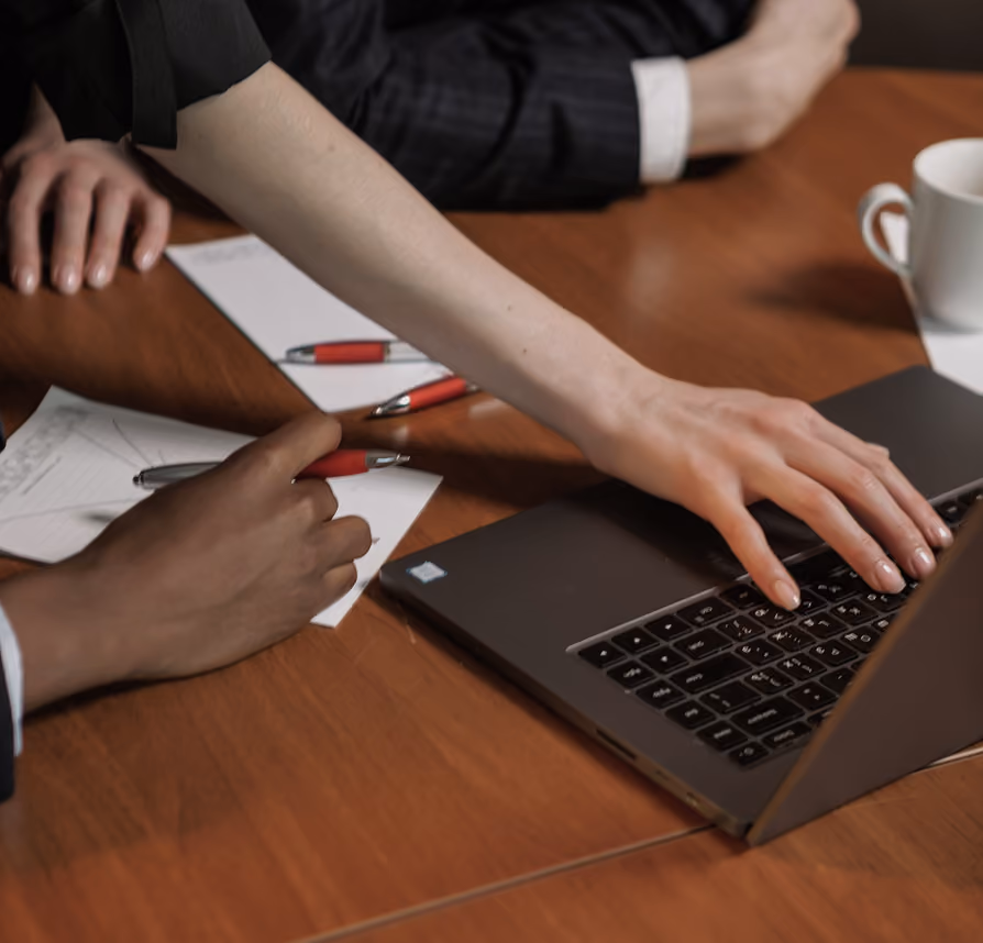 Two people at a table working with a laptop, pens, papers, and a coffee cup.