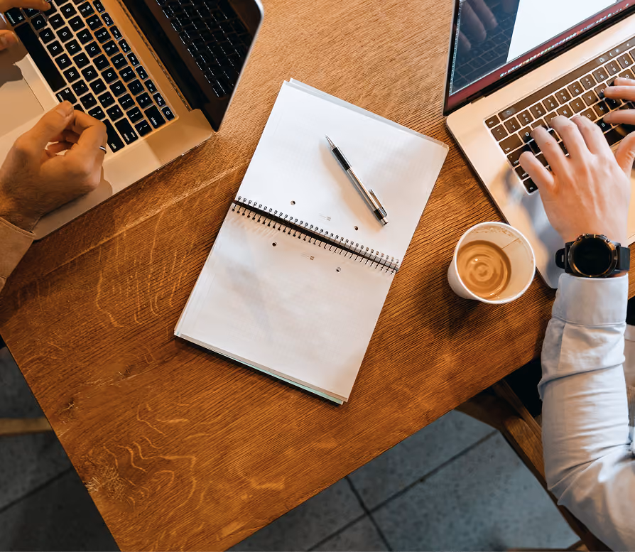 Two people typing on laptops at a wooden table with a notebook, pen, and a cup of coffee.