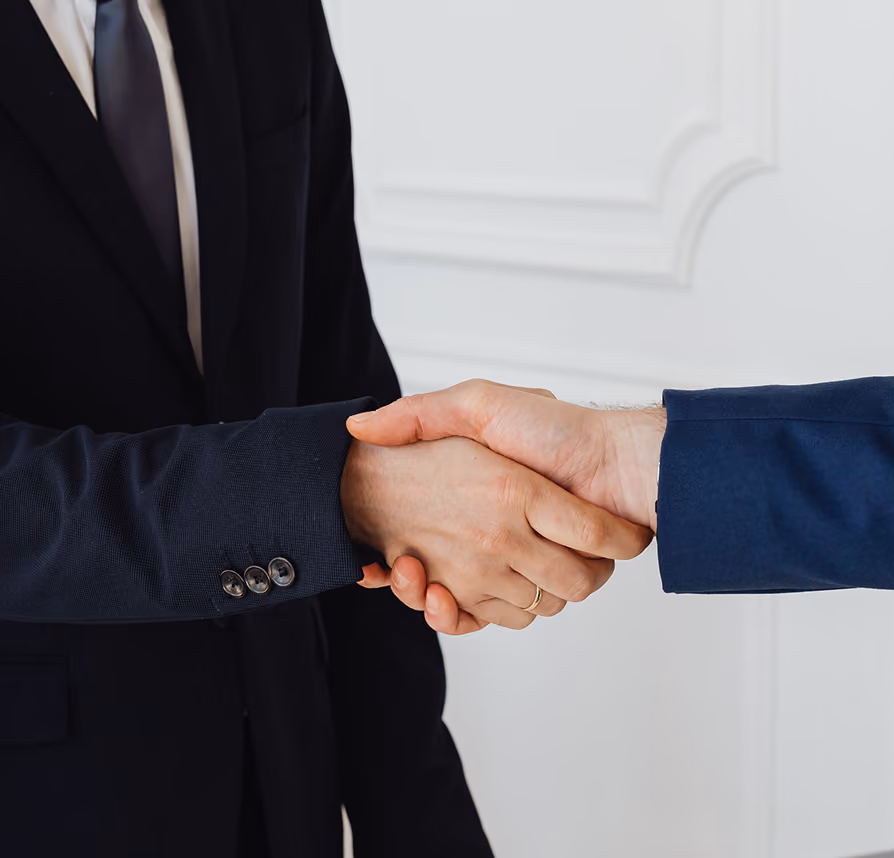 Two men in suits shaking hands in a formal setting with white decorative wall background.