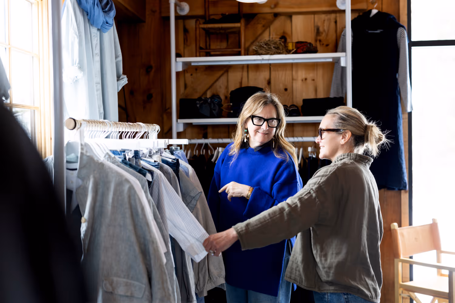 Two women browse and discuss clothes on a rack inside a cozy wooden boutique.