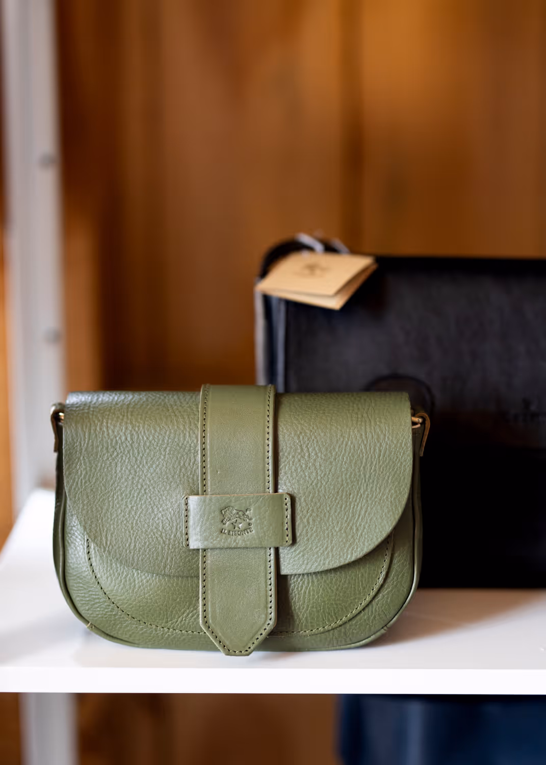 Green leather shoulder bag with front strap closure on a white surface, wooden background.