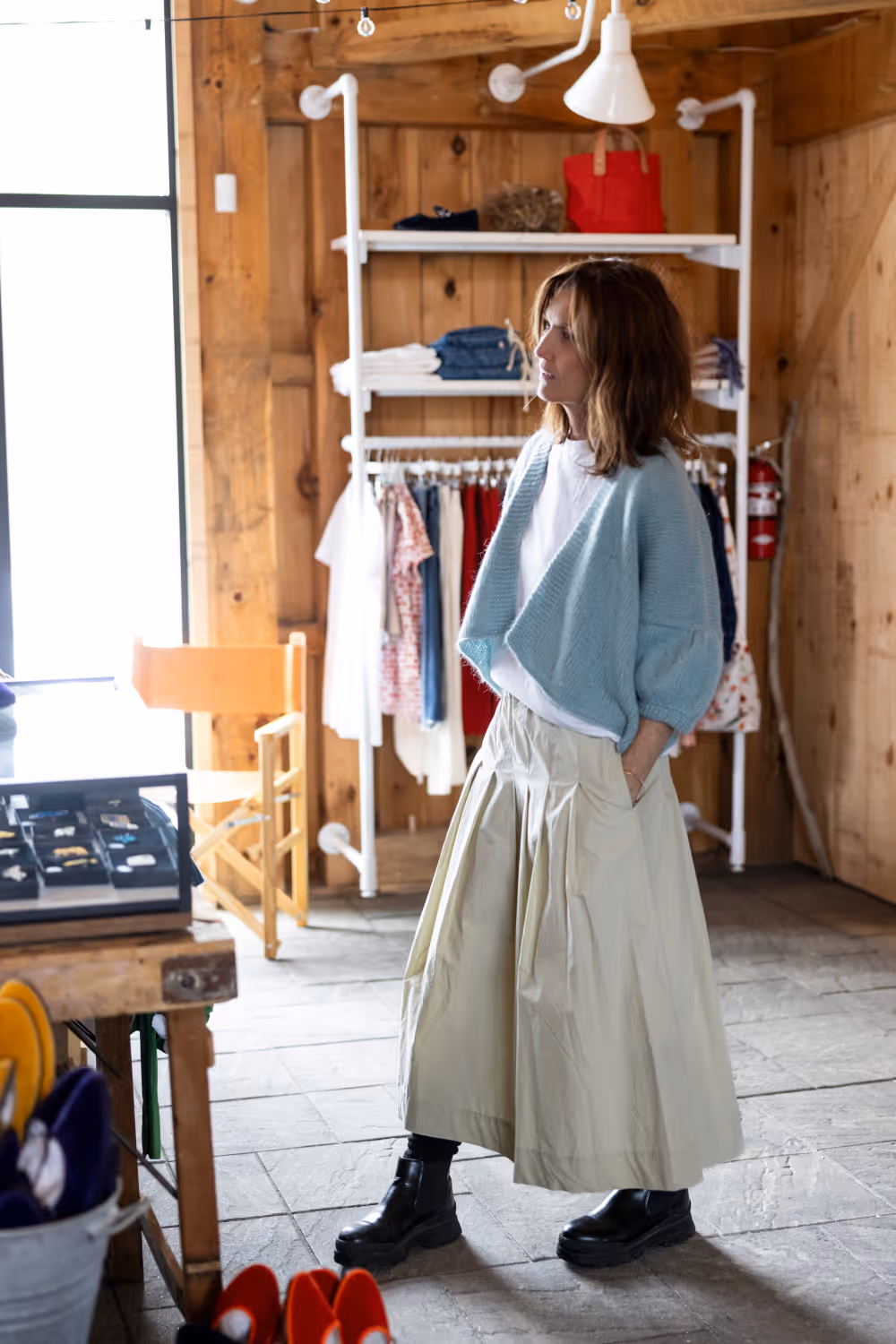 Woman in a blue cardigan, white shirt, beige skirt, and black boots standing in a wooden clothing store.