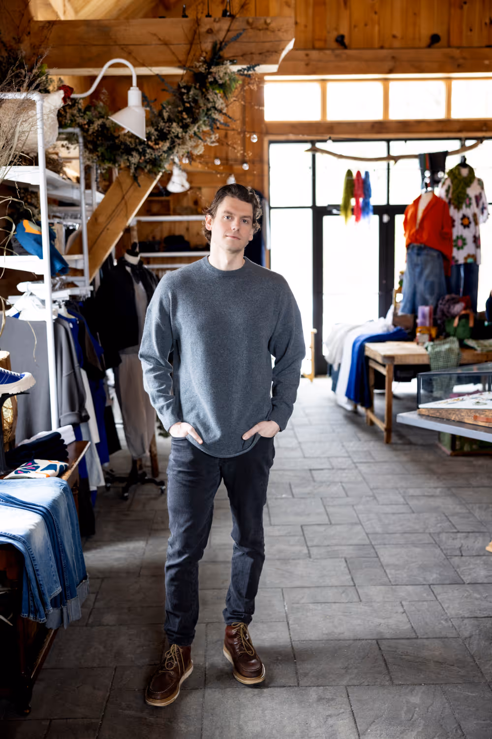 Man in gray sweater and dark pants standing in a boutique clothing store with wooden walls.