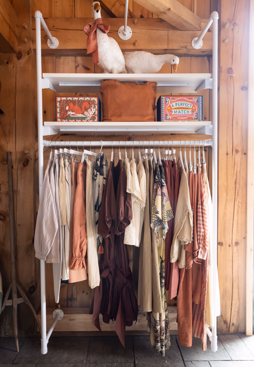 Clothing rack with neutral and rust-colored clothes under shelves holding two white ceramic geese and decorative boxes.