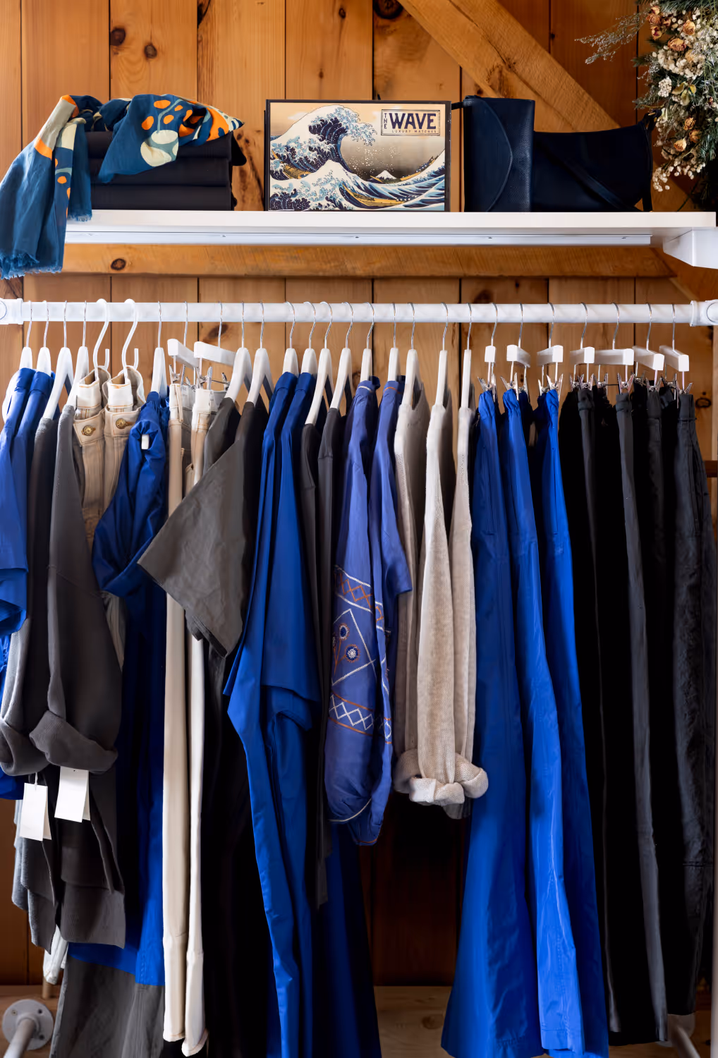 Clothes rack with blue, gray, beige, and black pants and shirts hanging on white hangers against a wooden wall.