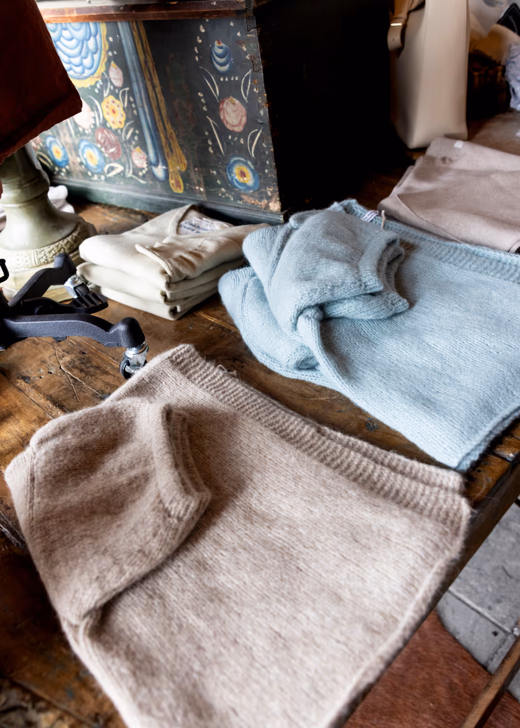 Folded neutral and light blue knit sweaters and tops on a rustic wooden table in a cozy setting.