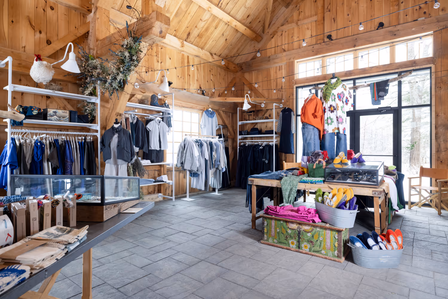 Bright boutique with wooden walls, clothing racks, a table with shoes and folded apparel in a rustic setting.