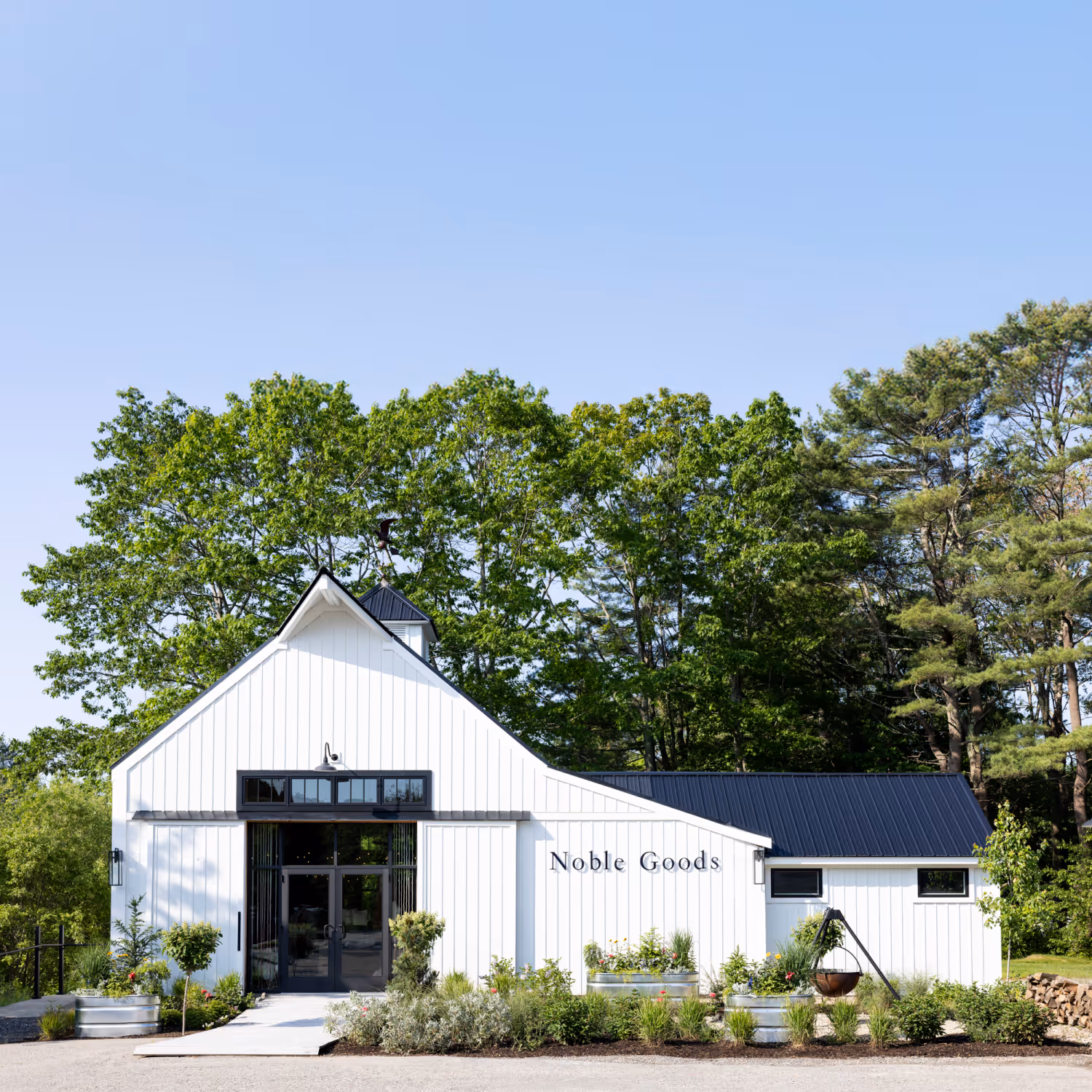 White building with black roof and glass doors labeled Noble Goods surrounded by green trees and plants.